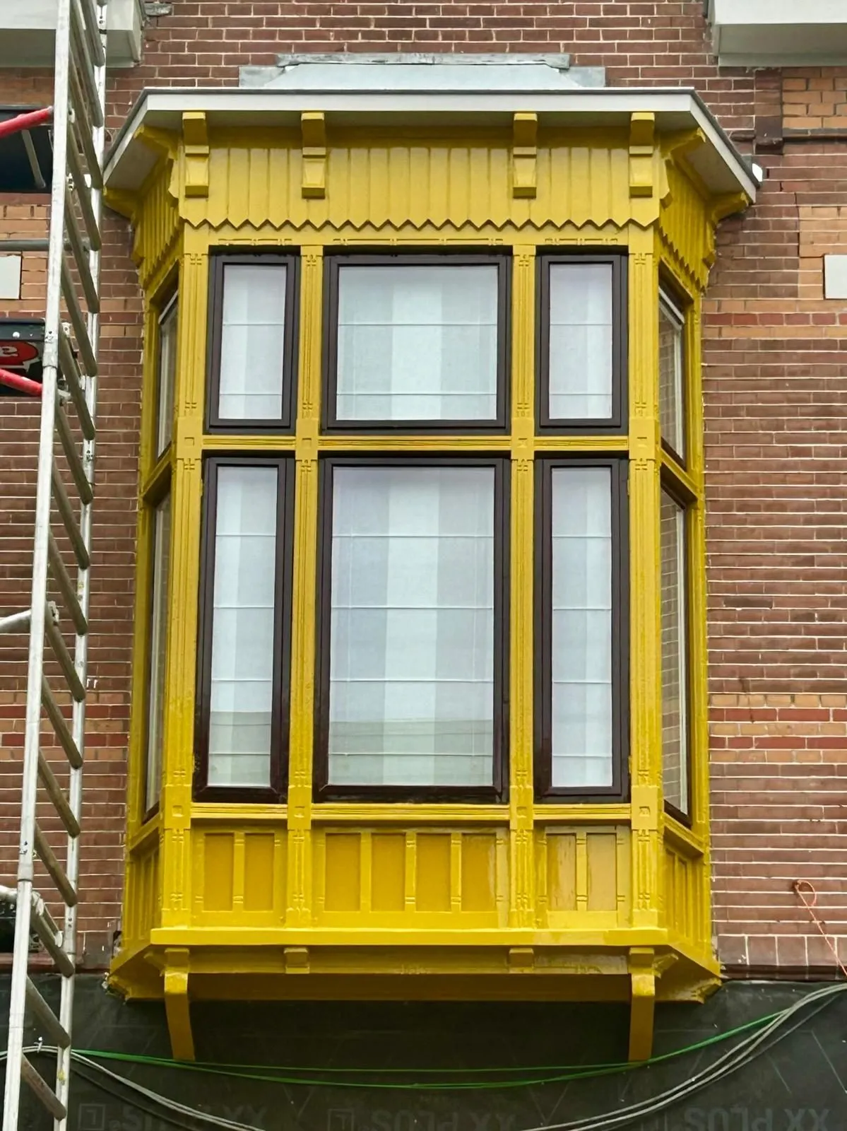 Houten erker in warm okergeel aan een bakstenen gevel