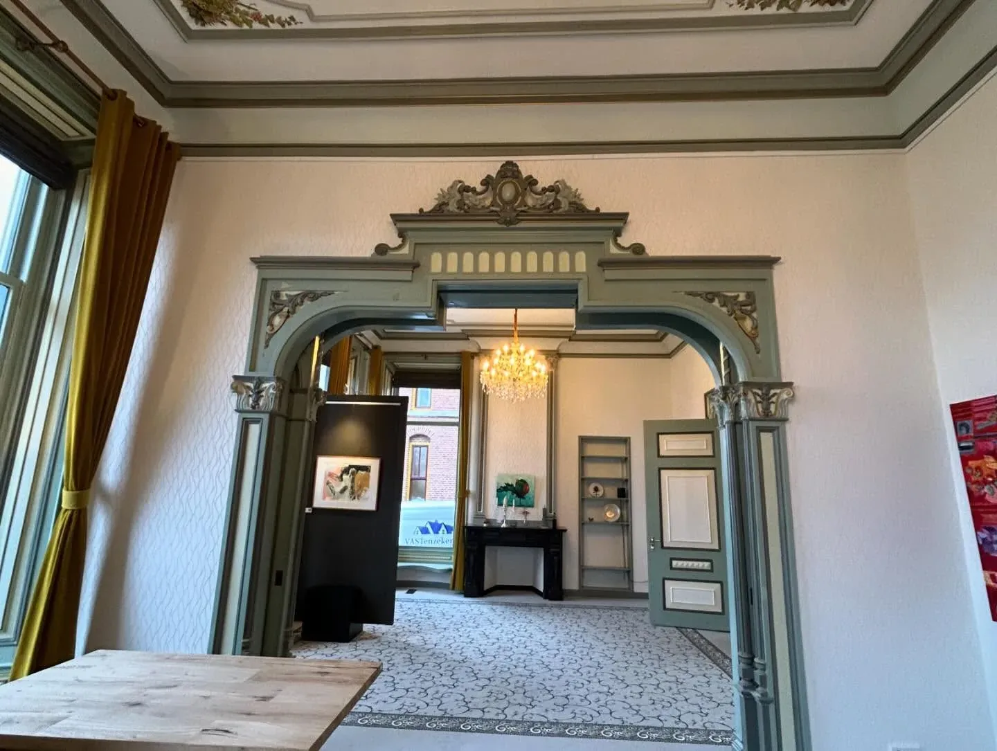 Doorkijk door een ornamentboog met vergulde details naar een zaal met kroonluchter