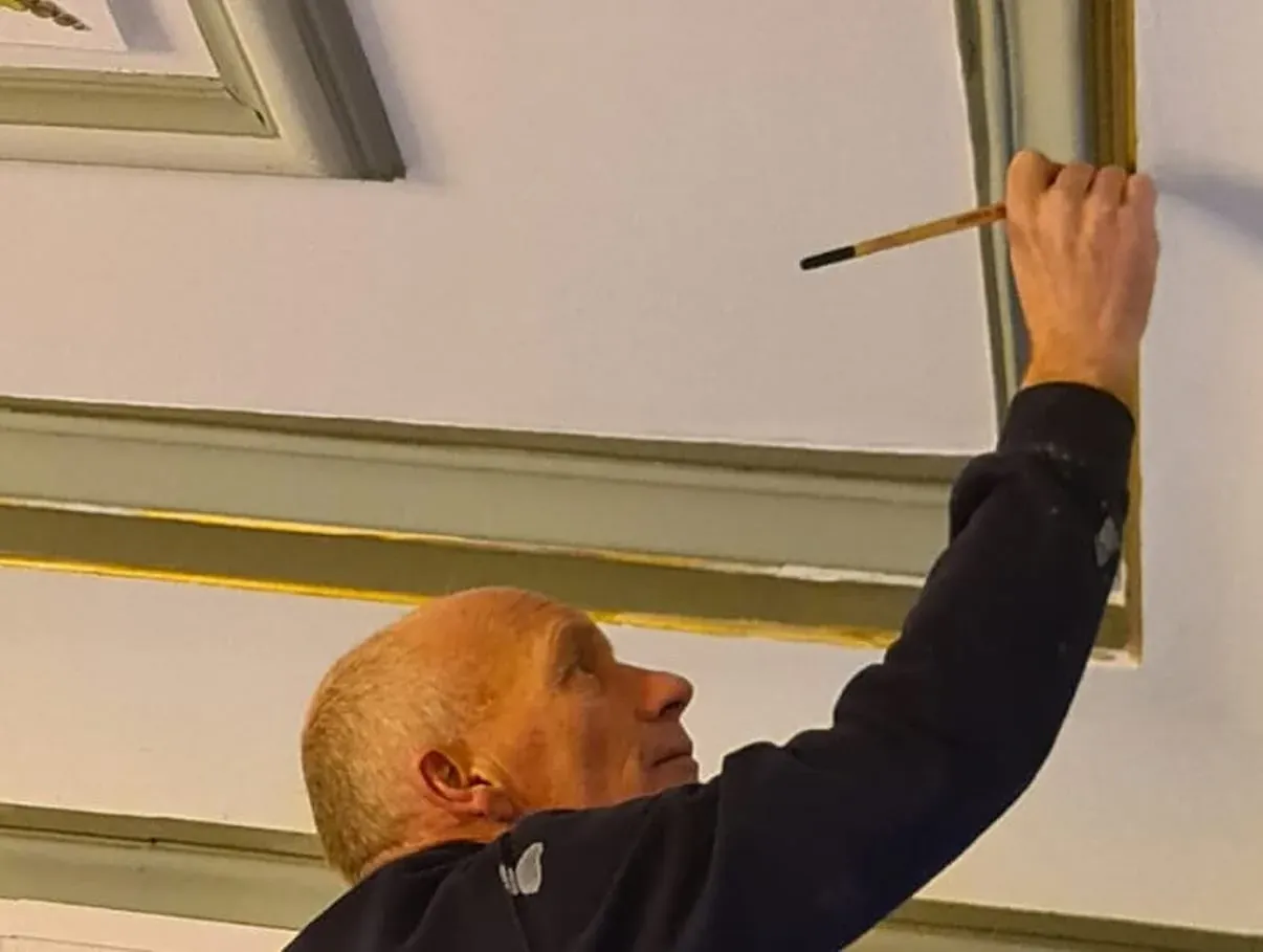 Een hand met een fijne kwast trekt een lijn langs het lijstwerk van een geschilderd plafond