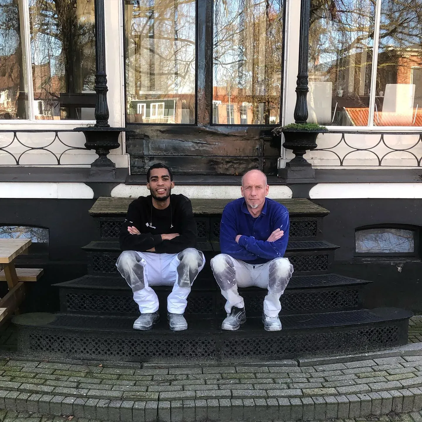 Twee schilders zitten op het bordes van hetzelfde pand, met het serreraam achter zich