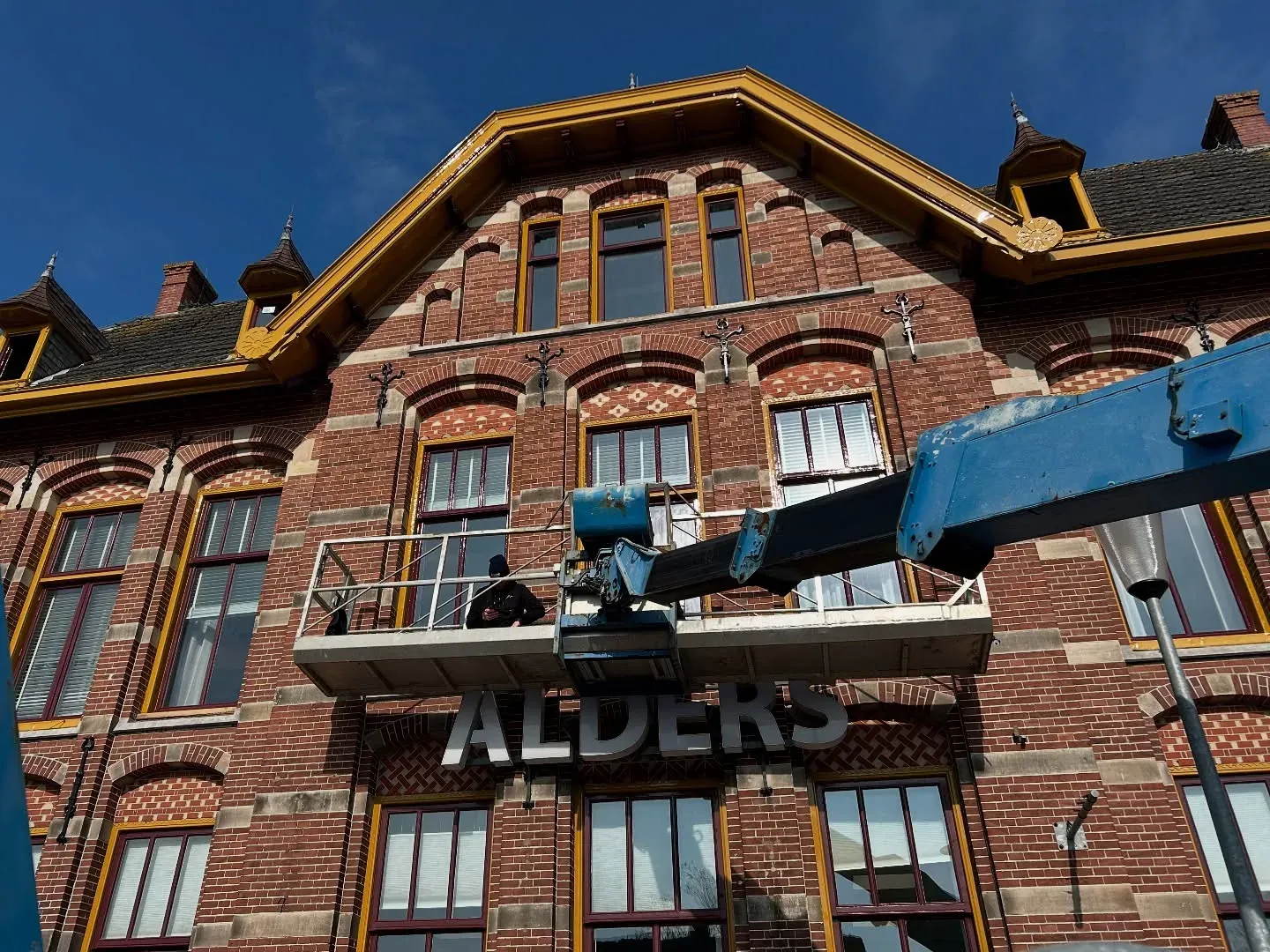 Bakstenen gevel met houten kapitelen en gele kozijnen, met een hoogwerker ervoor